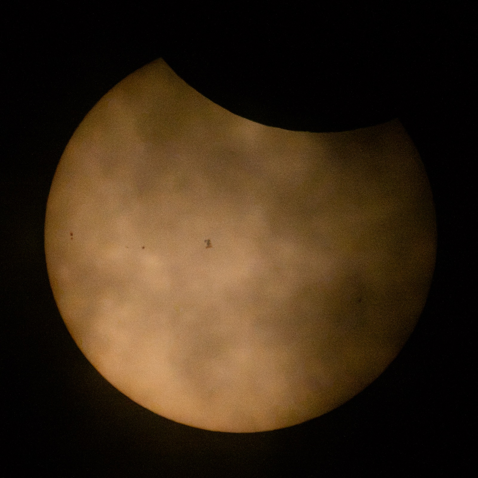 The International Space Station as a small dark silhouette on the face of the Sun, with the Moon covering the upper edge, photographed from the ground on 12 August 2026.